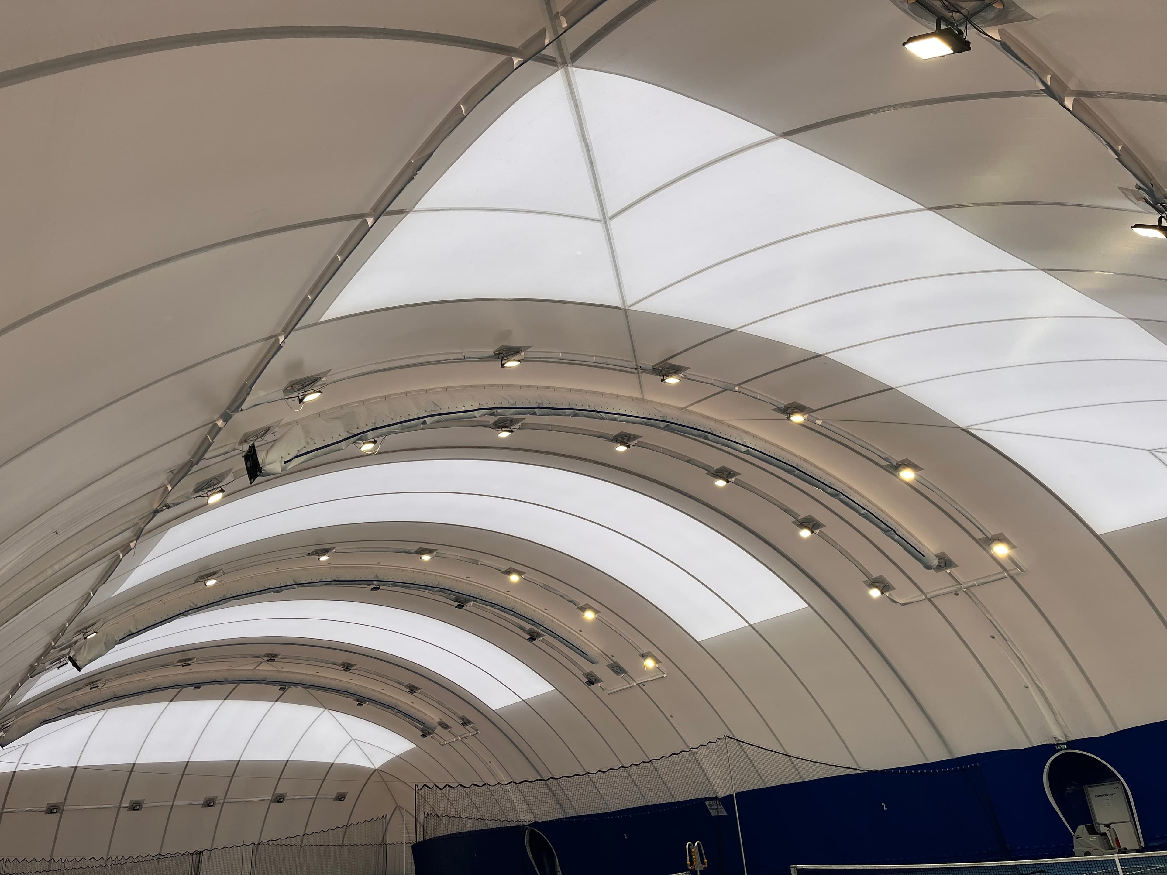 Arched white ceiling of a sports dome with rows of lights and translucent fabric panels.