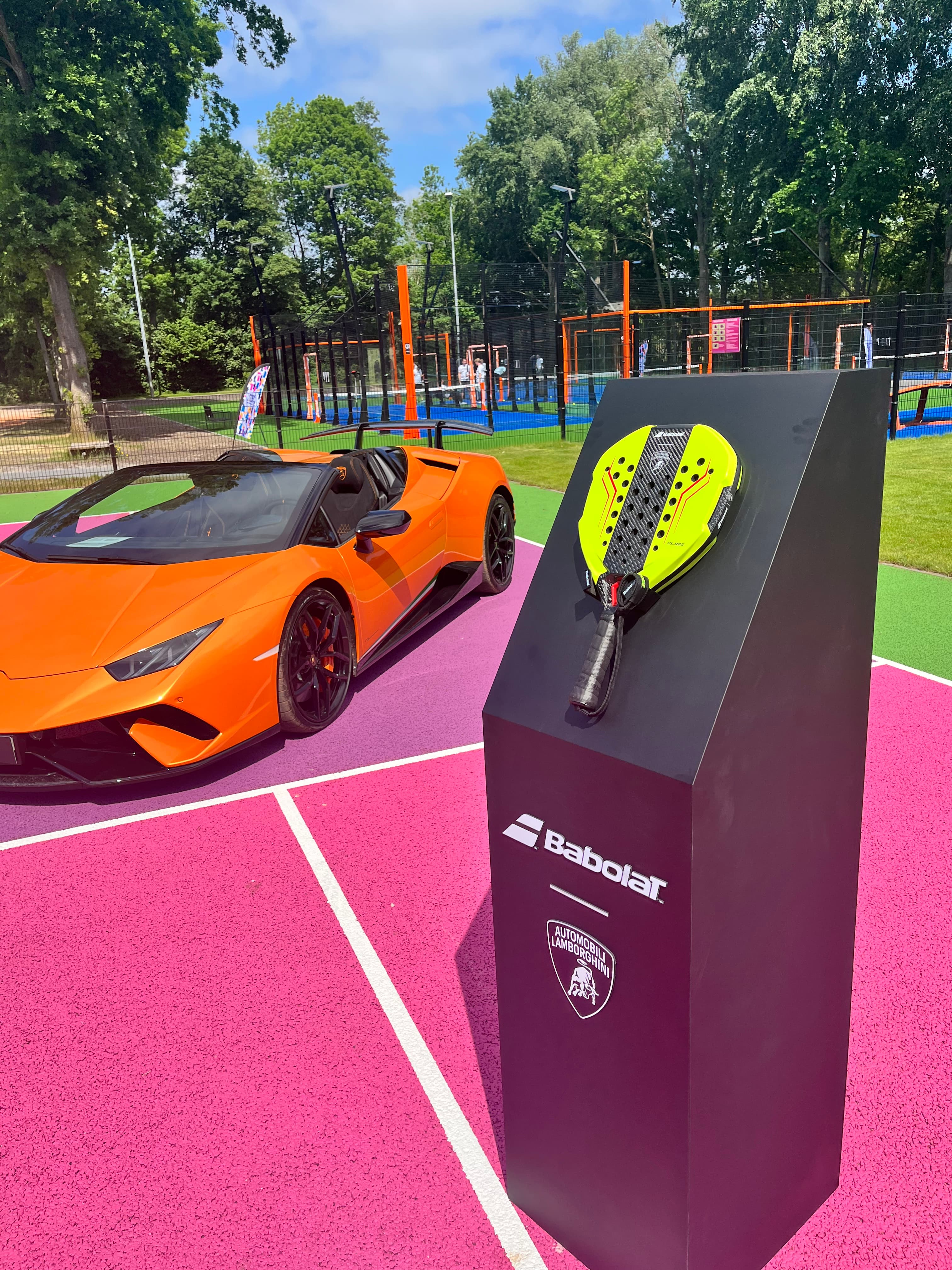 Orange Lamborghini sports car and yellow Babolat padel racket on a vibrant pink court.