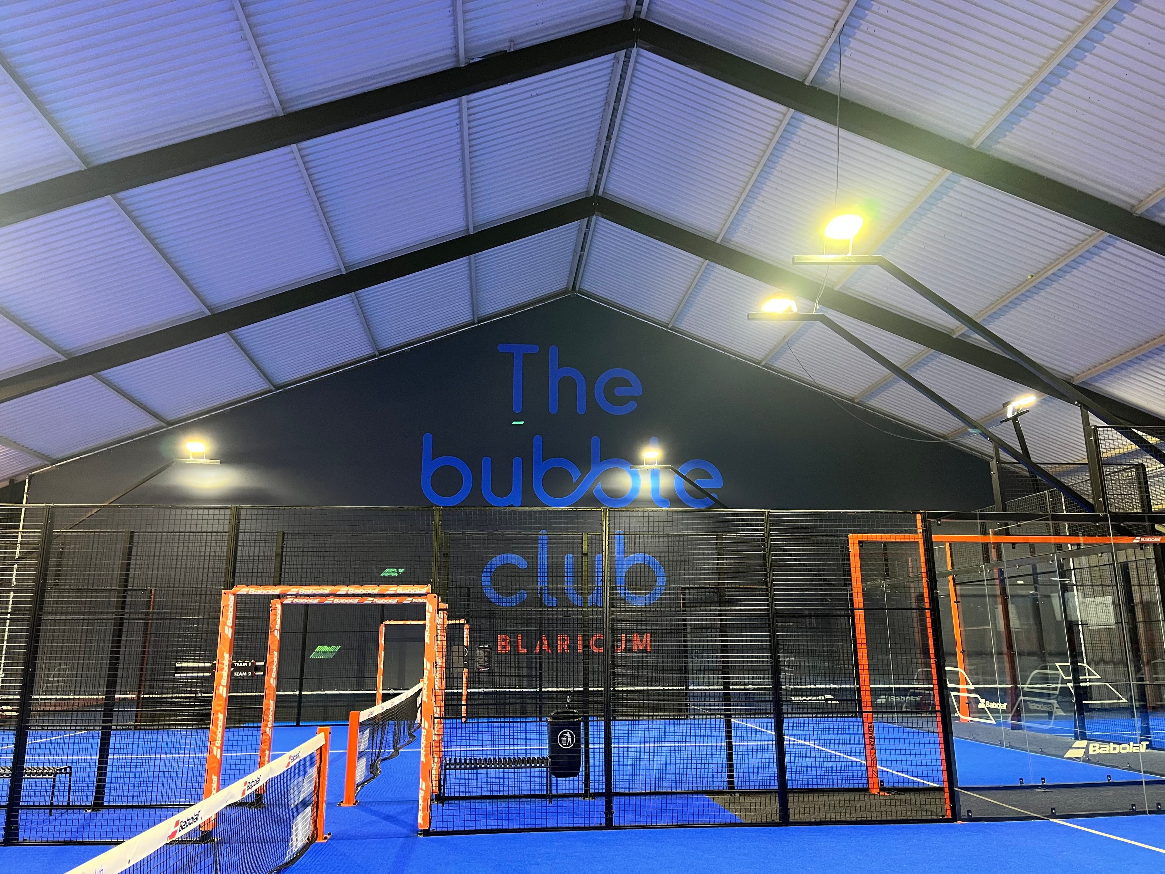 Indoor padel court with blue turf, black fencing, and 'The bubble club' wall signage.