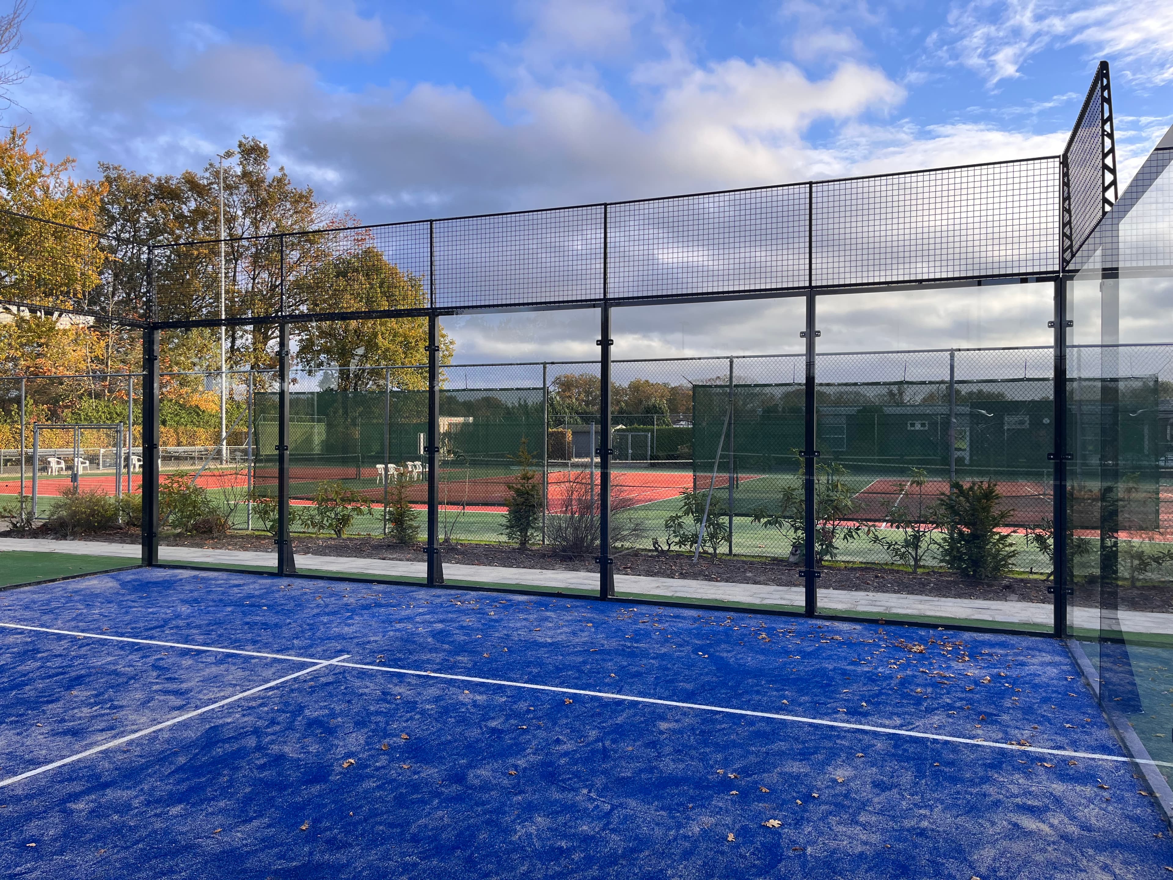 Blue padel court with glass walls, overlooking red clay tennis courts and autumn foliage.