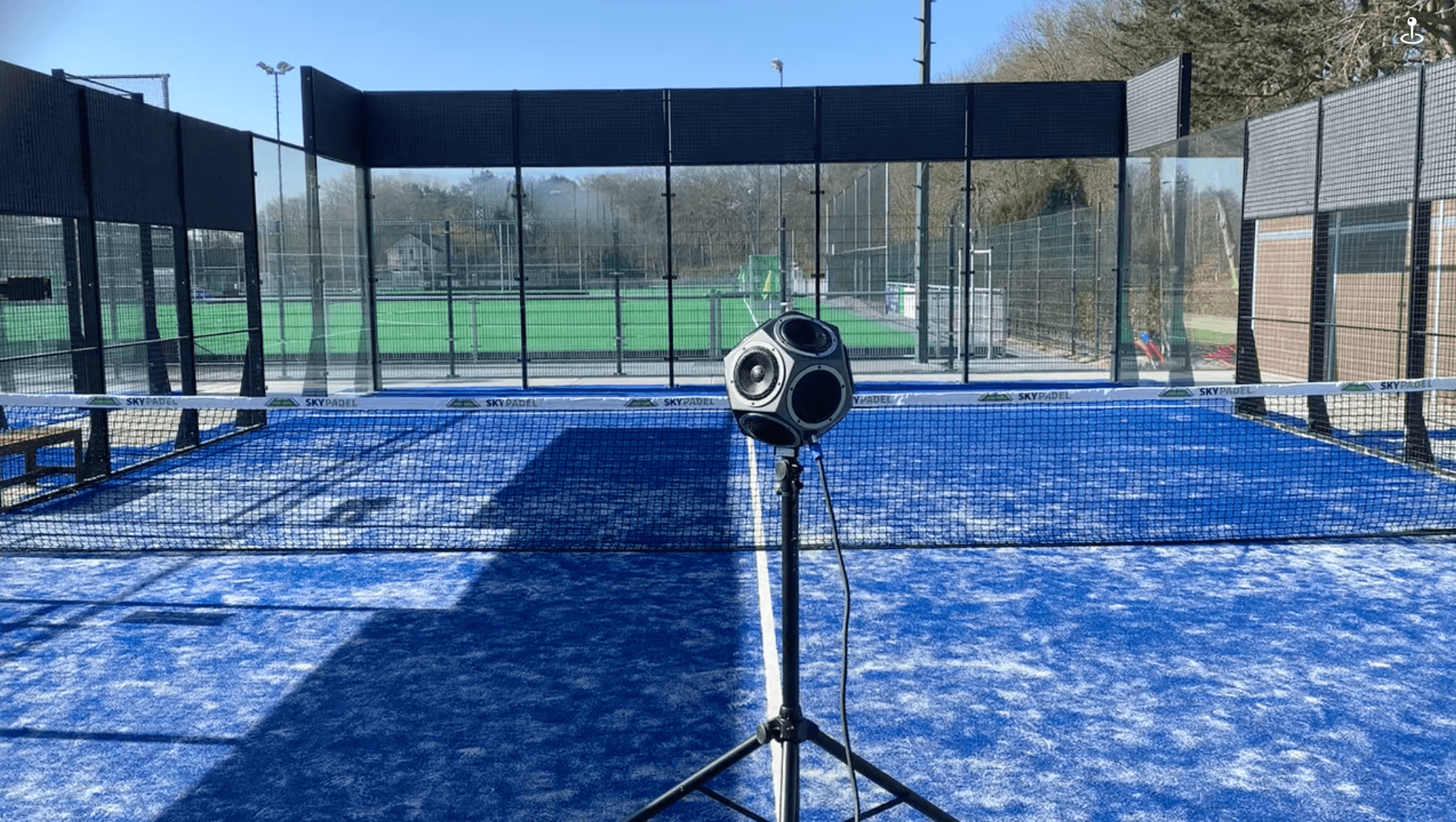 Spherical speaker on a tripod positioned in the middle of a blue outdoor padel court.