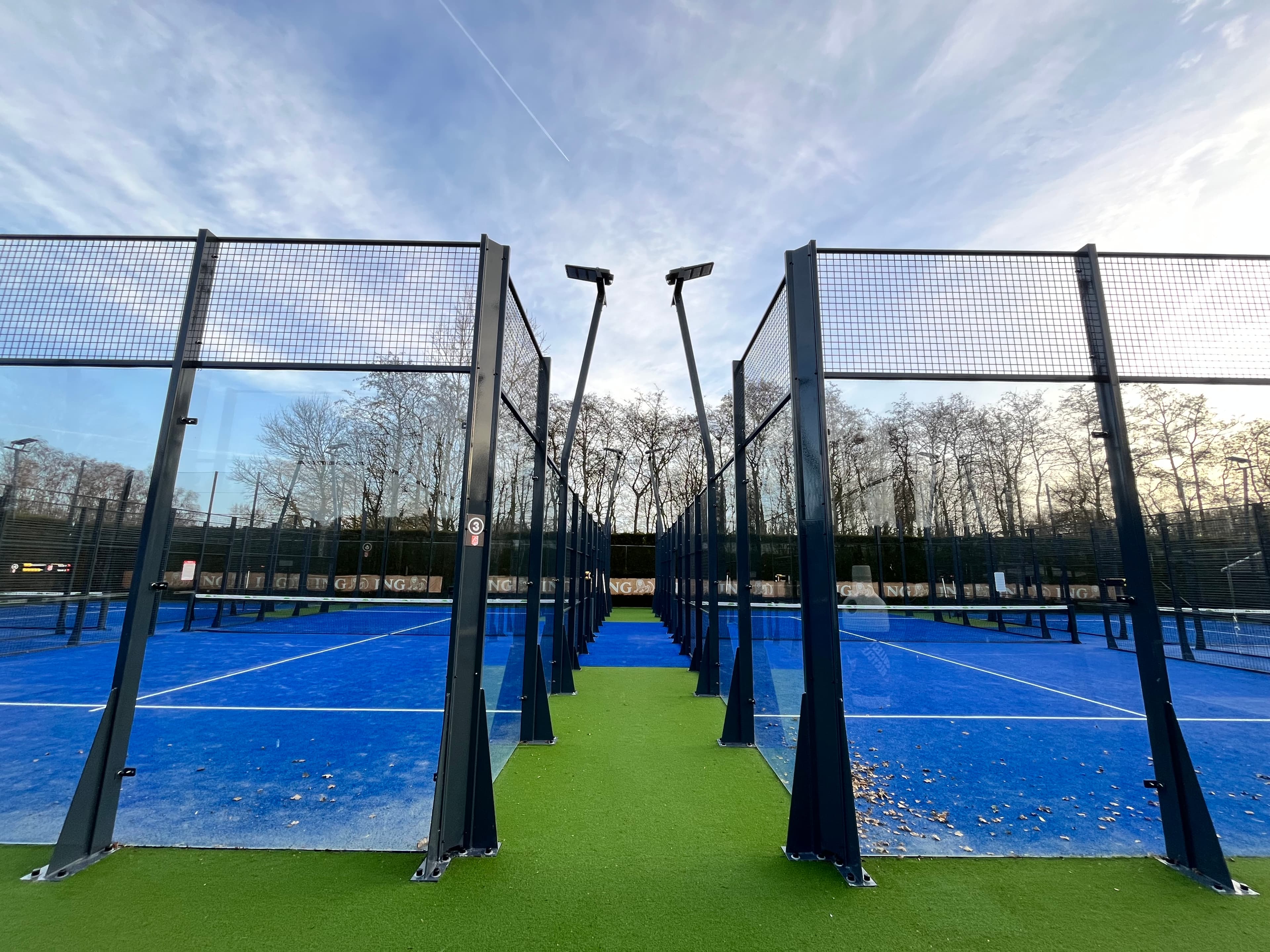 Blue padel courts with glass walls and metal fencing along a green turf path.