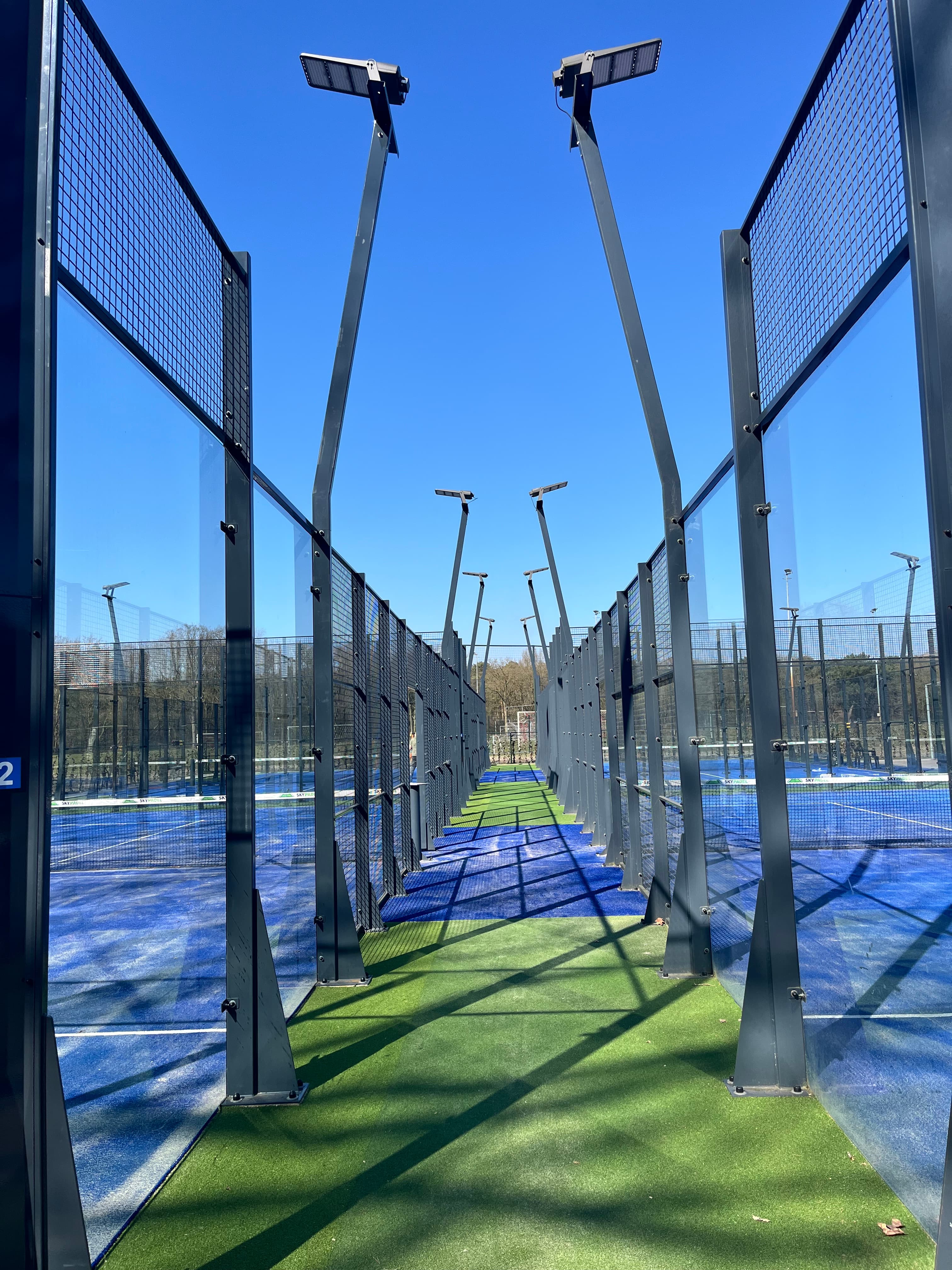 Padel courts with blue and green turf, glass walls, and floodlights under blue sky.