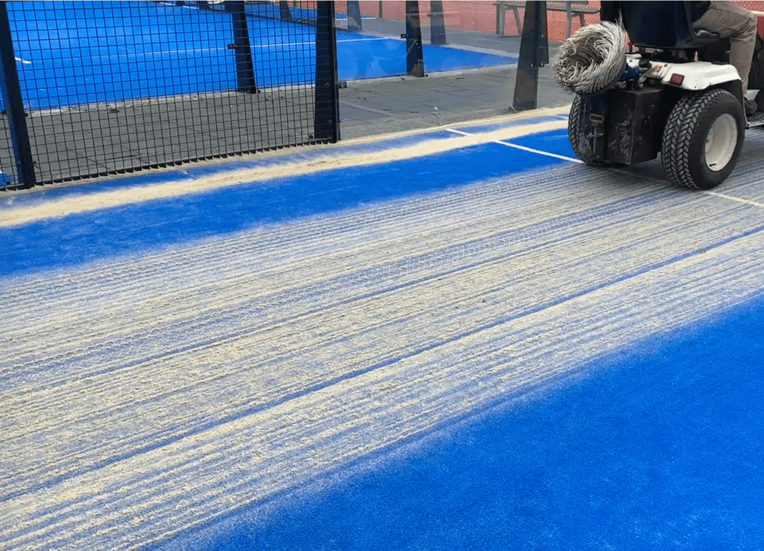 A motorized maintenance machine brushes sand onto a bright blue sports court surface.