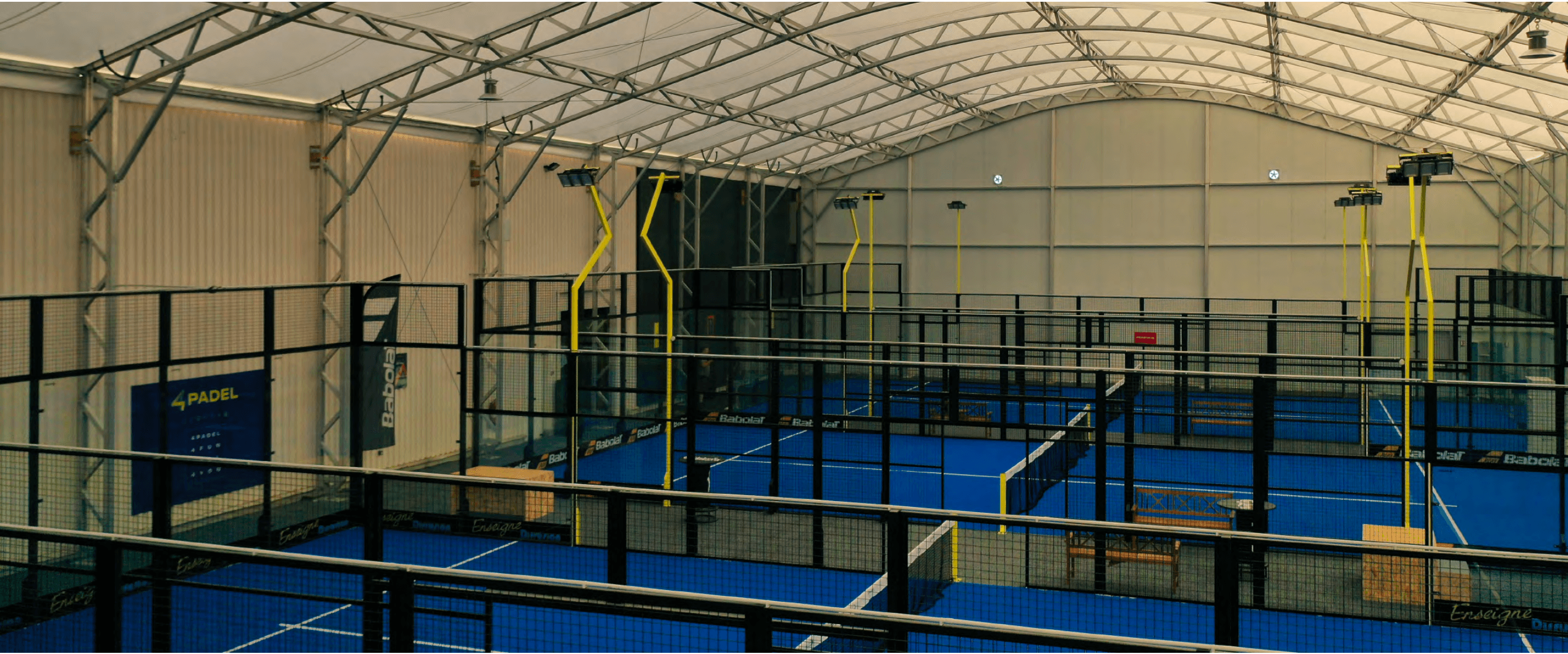 Multiple blue indoor padel courts with black fencing and yellow lights under an arched roof.