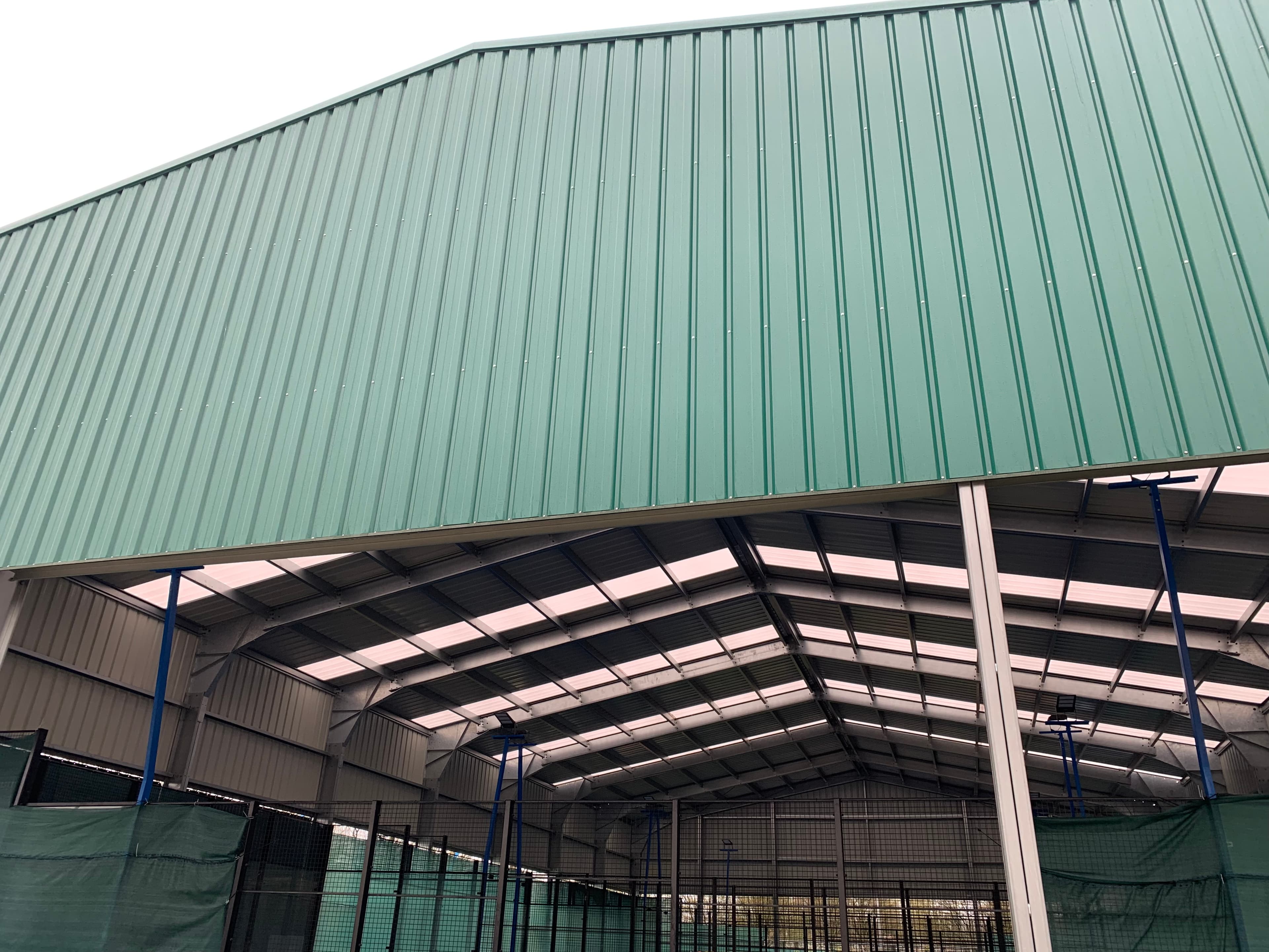 Large green corrugated metal sports hall with a visible interior featuring skylights and fencing.