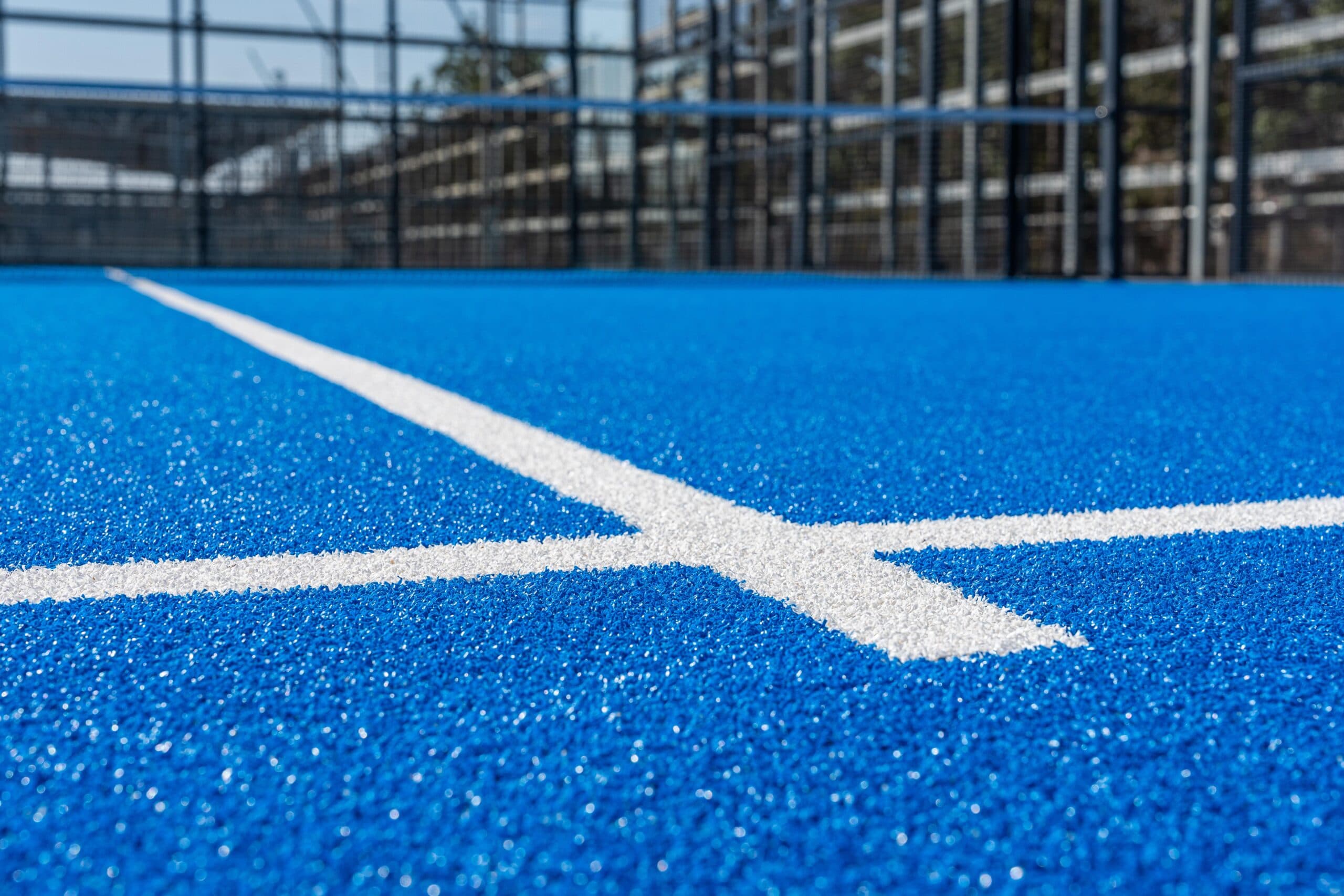 Close-up of white lines on a blue padel court with blurred fencing in the background.