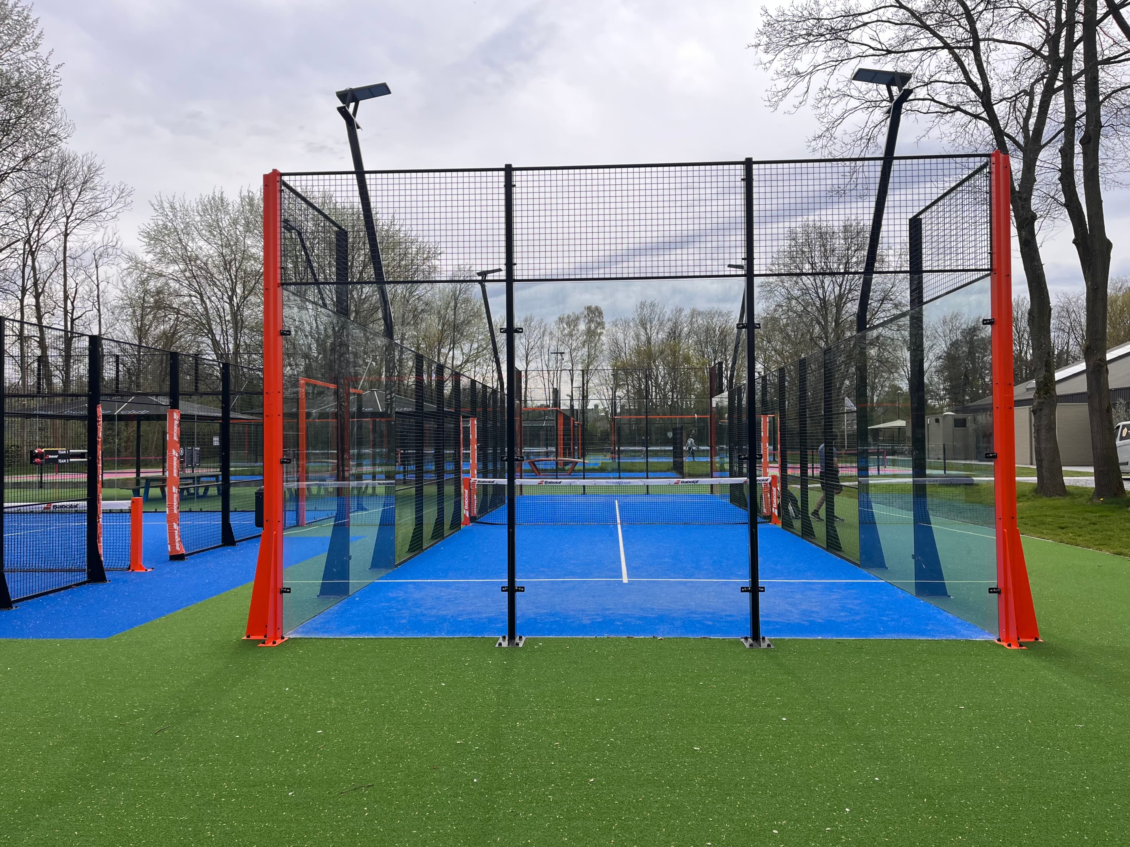 Outdoor padel court with blue turf, glass walls, and orange frames under an overcast sky.