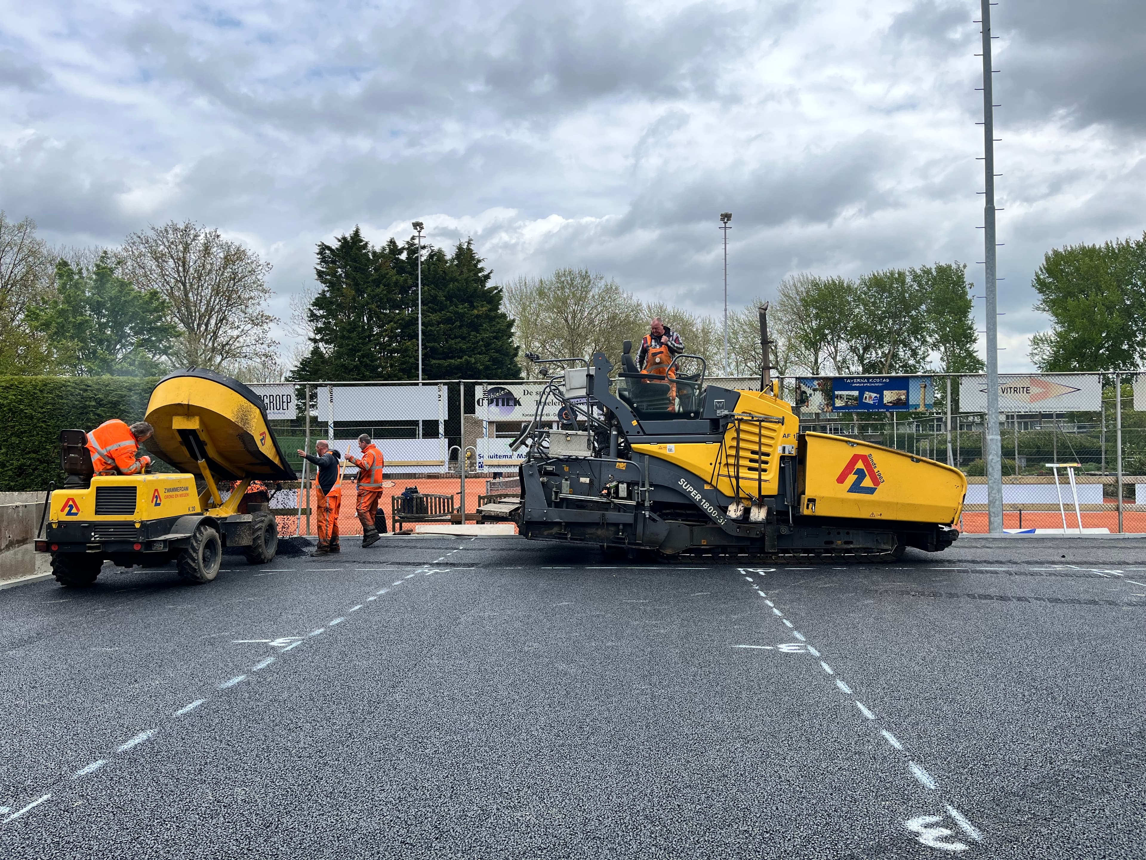 Workers in orange safety gear use a yellow asphalt paver and dumper to lay pavement.