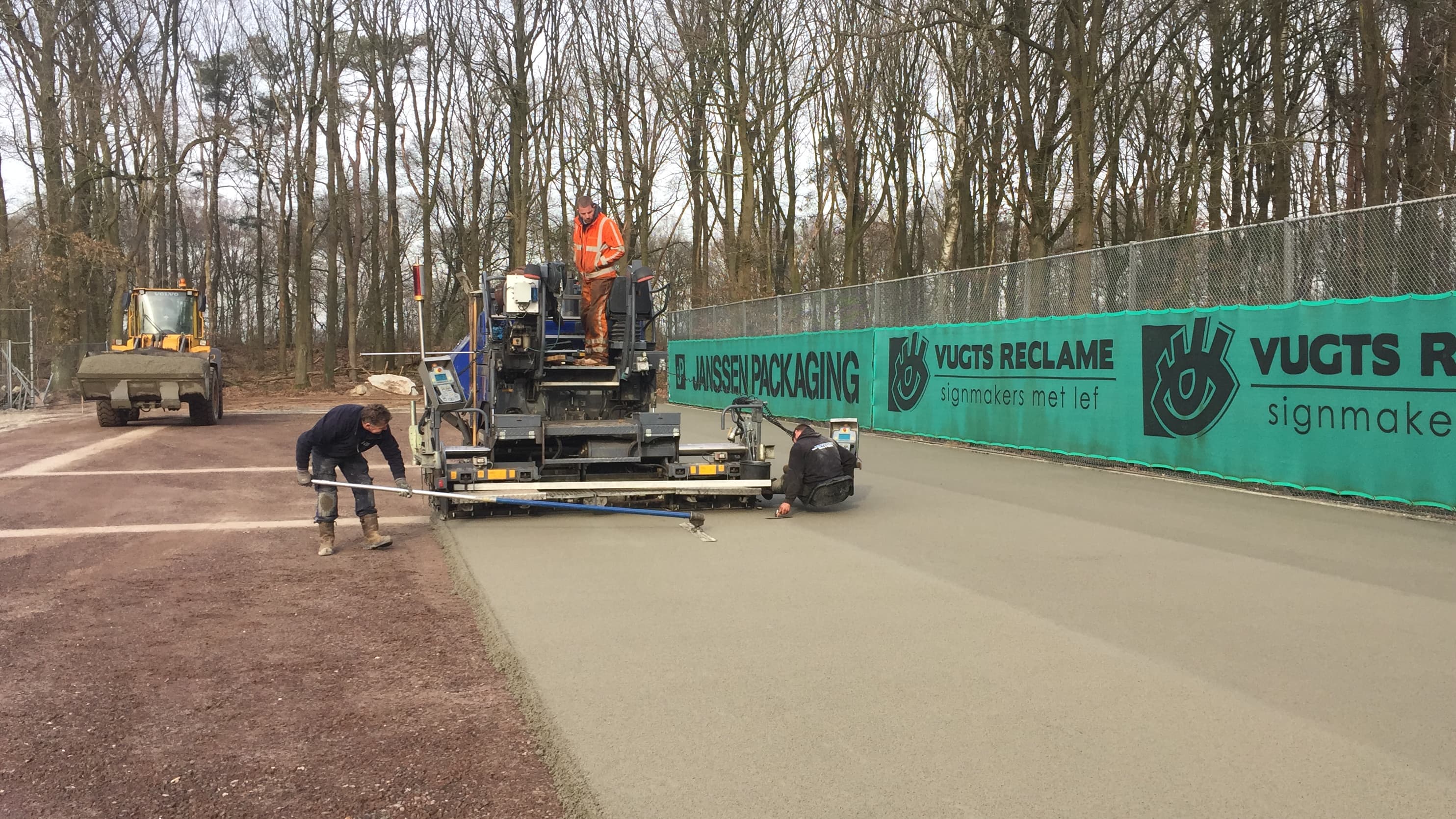 Workers using a paving machine to lay a smooth concrete surface next to a fence.