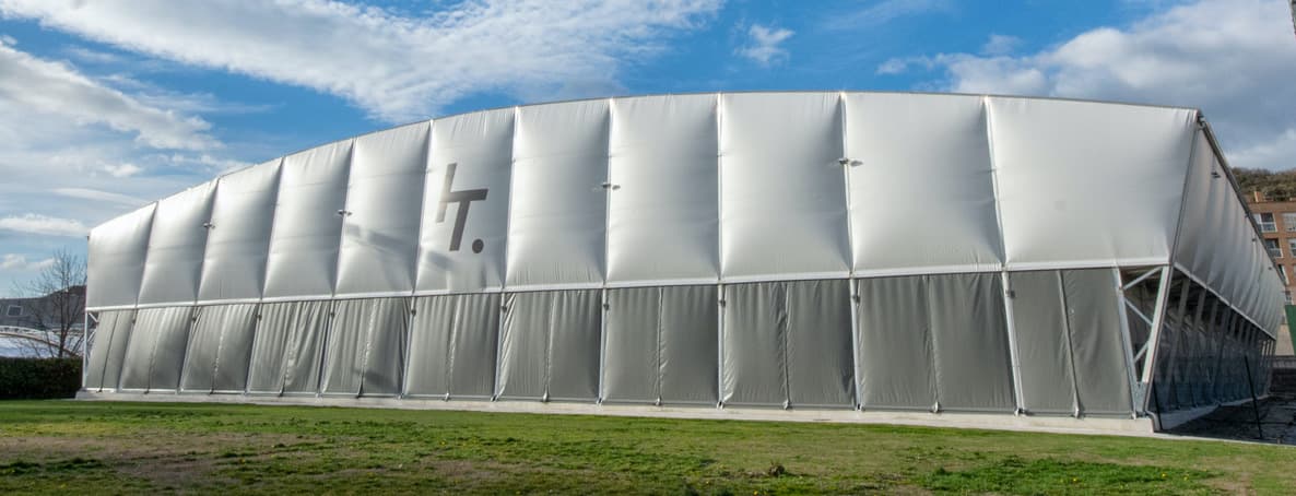 Large white fabric sports hall with grey panels on a grassy field under blue sky.